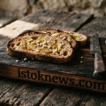 Несколько ломтиков поджаренного черного хлеба с чесноком, лежащих на деревянной доске рядом с головкой чеснока.