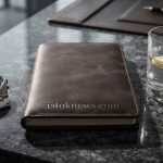 Строгий мужской натюрморт: кожаный блокнот, элитные часы и стакан воды на темном фоне.