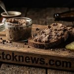 Стеклянная баночка с густой белой пастой, в которой видны кусочки зелени, рядом лежат ломтики черного хлеба с толстым слоем намазки.