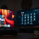 Игровая консоль возле включенного экрана телевизора в вечерней комнате